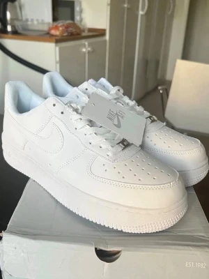 Nike Air Force 1 vita sneakers - Klassiska Nike Air Force 1 sneakers i helvitt läder med perforerad tå, vit sula och vita skosnören. Ikonisk swoosh på sidan och metallbricka på snörningen. Stilren och tidlös modell som funkar till det mesta. Kommer med originalkartong och taggar.