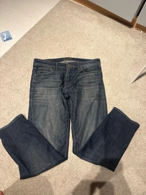 Blå raka jeans med patchade ben - Säljer ett par blå jeans med raka ben och patchade detaljer på baksidan av benen. Jeansen har klassisk femficksdesign, knappgylf och en läderpatch bak i midjan. Materialet är denim i bomull och färgen är tvättad blå med slitningar.