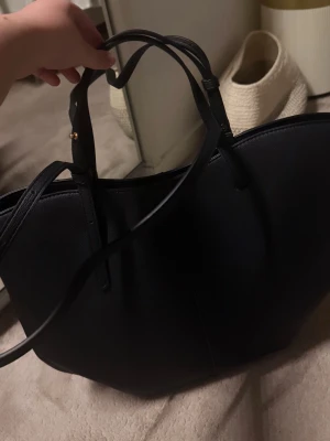 Svart shoppingväska  - Stor och stilren svart shoppingväska med två handtag och en axelrem. Väskan har en enkel design med lätt struktur i materialet och guldfärgade detaljer vid handtagen. Perfekt för dig som gillar minimalistisk look och vill ha plats för allt. Ny köpt men säljer den då jag har en till! 