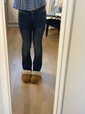 Mörkblå bootcut jeans - Säljer ett par mörkblå bootcut jeans med klassisk femficksdesign och lätt slitna detaljer. Jeansen har normal midja och är tillverkade i ett stretchigt denimtyg som sitter snyggt på benen. Perfekta till en avslappnad look.