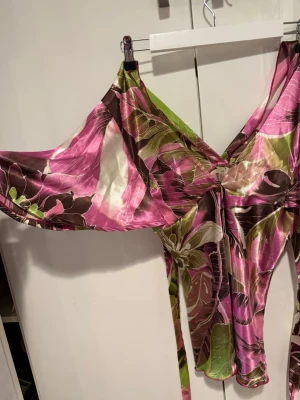 Blommig omlottklänning i satin - Säljer en färgstark omlottklänning i satin med blommigt mönster i rosa, grönt, brunt och vitt. Klänningen har v-ringning, vida ärmar och knyt i midjan för en snygg passform. Perfekt för dig som vill sticka ut med en unik look!