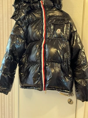 Svart glansig pufferjacka Moncler - Svart glansig pufferjacka från Moncler med huva och ikonisk logga på bröstfickan. Jackan har en röd, vit och blå randig dragkedja framtill, elastiska muddar och flera fickor. Tillverkad i blankt material som ger en cool look.