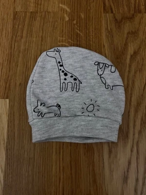 Grå barn / baby mössa med djurmotiv, storlek 0-2 månader - Fin grå babymössa med gulligt djurmotiv säljes! Motiv: Giraff, ko, hare och sol i svart tryck. Storlek: Storleken framgår inte, men jag uppskattar att den passar barn mellan ca 0–2 månader. Säljs i bra skick! Kika gärna in på mina andra annonser, jag säljer just nu flera barn- och babyplagg samt barnartiklar! Hör av dig vid intresse och frågor!