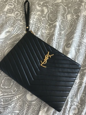 Svart kuvertväska från YSL i läder - Snygg svart kuvertväska från YSL i quiltat läder med guldigt YSL-emblem framtill. Väskan har ett stilrent mönster i v-format och en avtagbar handledsrem. Perfekt för att lyfta din outfit med en lyxig touch. En liten skavank se bild, men inget som märks vid användning annars är den i nyskick 