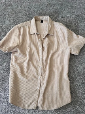 Beige manchesterskjorta från SHEIN - Kortärmad skjorta i beige manchester från SHEIN. Skjortan har klassisk krage, knappar framtill och en avslappnad passform. Perfekt för dig som gillar en enkel men ändå trendig stil.
