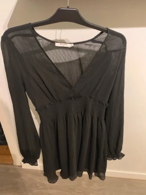 Svart omlottklänning från NA-KD - Snygg svart kortklänning från NA-KD med omlottdesign och v-ringning. Klänningen har långa, transparenta ärmar med resår vid handlederna och smock i midjan för en bekväm passform. Perfekt för dig som gillar en stilren och trendig look.