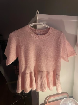 Ljusrosa stickad topp från Gina Tricot - Supersöt ljusrosa stickad topp från Gina Tricot med korta ärmar och volang nertill. Mjuk och fluffig känsla, rund halsringning och ribbstickade detaljer vid hals och volang. Perfekt för en gullig och trendig look.