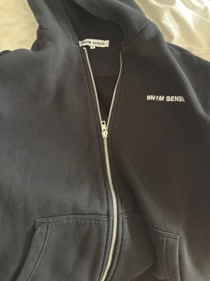 Svart hoodie-kappa från 9N1M SENSE - Svart hoodie-kappa från 9N1M SENSE med dragkedja framtill och två stora fickor. Klassisk huva och broderad logga på bröstet. Perfekt för en avslappnad streetwear-look. Tillverkad i mjukt bomullsmaterial.