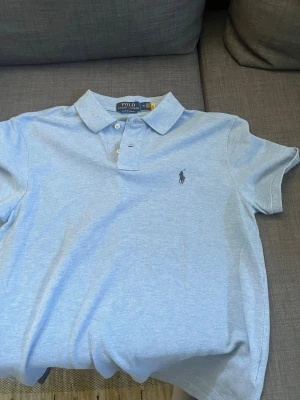 Ljusblå piké från Polo Ralph Lauren - Säljer en ljusblå pikétröja från Polo Ralph Lauren i custom slim fit. Klassisk krage, korta ärmar och den ikoniska broderade loggan på bröstet. Perfekt till sommaren och enkel att matcha med jeans eller shorts. Riktigt snygg och är perfekt till sommaren eller bara allmänt trevlig och skön att bära. Pris kan alltid diskuteras vid snabb affär. Hör av dig vid minsta fundering!!
