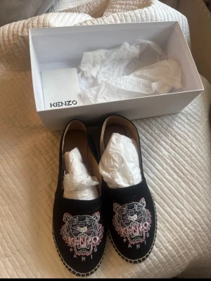 Svarta Kenzo loafers med tigerbrodyr - Säljer ett par svarta loafers från Kenzo med ikonisk tigerbrodyr i rosa, vitt och grått framtill samt texten 'KENZO PARIS'. Skorna har en klassisk slip-on modell med kontrastsömmar runt sulan och gummisula. Perfekta för dig som vill sticka ut med en unik detalj.