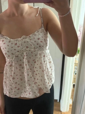 Vit blommig linnetopp - Söt vit linnetopp med små rosa blommor och tunna axelband. Toppen har en volangkant längs bysten. Luftigt material som passar perfekt till sommaren.