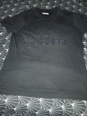 Svart Lacoste t-shirt med logga - Svart t-shirt från Lacoste med stor broderad logga och text på bröstet. Klassisk passform och rund halsringning. Perfekt till jeans eller shorts för en clean och stilren look.