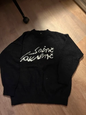 Svart Saint Laurent stickad tröja - Säljer en svart stickad tröja från Saint Laurent med vit logotyp i handskriven stil på bröstet. Tröjan har rund halsringning och långa ärmar. Perfekt för dig som gillar exklusiva och trendiga plagg.