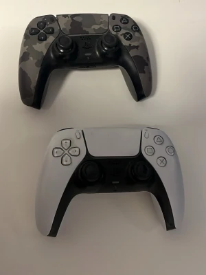 PlayStation 5 DualSense handkontroller båda har stick drift fråga om ni inte vet vad de e - Två PS5 DualSense handkontroller, en i svart och grått camomönster och en i klassiskt vitt med svarta detaljer. Ergonomisk design med responsiva knappar och triggers. Perfekt för gaming på PlayStation 5. Tillverkade i slitstark plast.