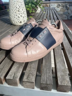 Valentino Garavani Open sneakers ljusbeige/blå - Snygga sneakers från Valentino Garavani i ljusbeige läder med bred mörkblå detalj över mitten. Helt äkta med bevis som kan skickas, Klassisk låg modell med matchande ljusbeigea snören och diskret logga på plösen. Perfekta för dig som vill ha en stilren men ändå unik look. Tillverkad i Italien 🇮🇹