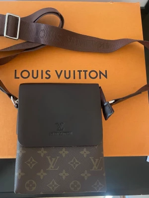 Lou Vu brun axelväska - Snygg axelväska från LouVu i klassiskt brunt monogrammönster. Väskan har ett lock i mörkbrunt läder, justerbar axelrem och en praktisk dragkedjeficka på baksidan. Perfekt för dig som vill ha en stilren och lyxig accessoar.