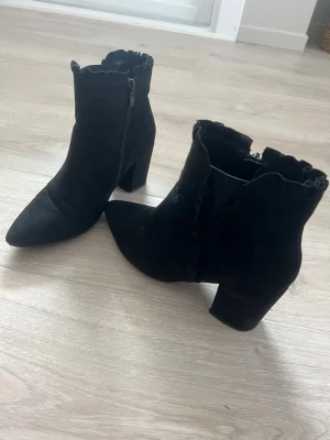 Svarta stövletter med volangkant - Säljer ett par svarta stövletter i mockaimitation med spetsig tå och chunky klack. Skorna har dragkedja på sidan och en snygg volangkant upptill som ger en extra detalj. Perfekta för dig som vill ha något stilrent men ändå unikt.