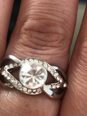 Silverfärgad ring med stor sten - Snygg silverfärgad ring med en stor, rund klar sten i mitten och små gnistrande stenar runt om. Ringen har en tvinnad design som ger en unik look. Perfekt för dig som gillar bling och vill sticka ut. 