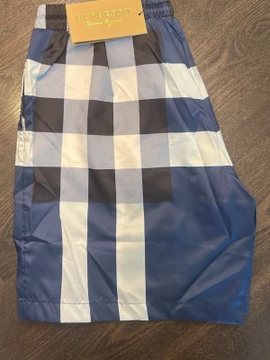 Burberry badbyxor  - Snygga badbyxor från Burberry med klassiskt rutmönster i blått, vitt och svart samt en diskret röd linje. De har elastisk midja och en bakficka. Materialet är snabbtorkande och perfekt för stranden eller poolen. Top kvale och använda en gång då de inte passade 