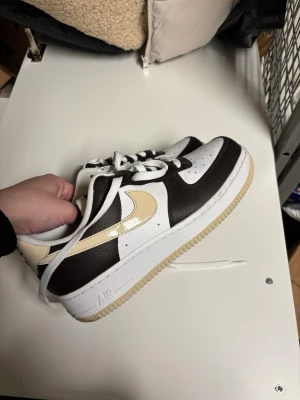 Nike Air Force 1 låga sneakers brun/vit - Nike Air Force 1 sneakers i lågt utförande med vit tå och sidor, mörkbruna paneler och beige swoosh. Ovandelen är i läder och sulan är vit med beige yttersula. Klassisk snörning och perforerad tå för ventilation.