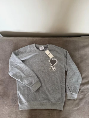 Grå sweatshirt från Ami Paris - Grå sweatshirt från Ami Paris med rund halsringning och ribbade muddar. Stor broderad logga med hjärta och bokstaven A på bröstet. Mjuk och bekväm, perfekt för en avslappnad stil.
