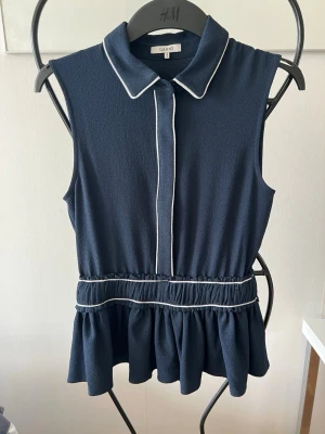 Mörkblå ärmlös topp från Ganni - Säljer en mörkblå ärmlös topp från Ganni med vit piping längs krage och knappslå. Toppen har en markerad midja med volangkant nedtill och klassisk skjortkrage. Perfekt för dig som gillar stilrena detaljer och en feminin siluett.