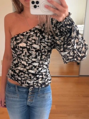 Svart och vit oneshoulder topp - Trendig oneshoulder topp med svart och vitt abstrakt mönster. Toppen har en puffig lång ärm på ena sidan och är åtsittande i midjan. Perfekt för dig som vill sticka ut med en unik och modern look. Strl m på lappen men insydd till en xs 