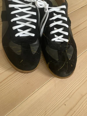 Svarta sneakers med mockadetaljer - Säljer ett par svarta sneakers med vita snören och gummisula. Skorna har både mocka och skinnpaneler samt diskreta gula stänk på ovandelen. Klassisk siluett som passar till det mesta och ger en clean look. Dok lite defekt 