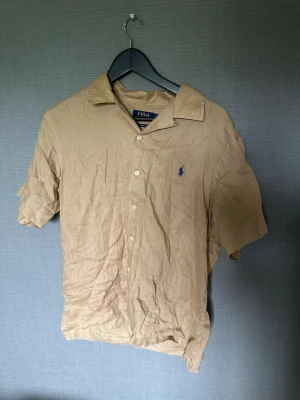 Beige linneskjorta från Polo Ralph Lauren - Snygg beige skjorta från Polo Ralph Lauren i linne med korta ärmar och klassisk krage. Skjortan har knappar framtill och den ikoniska blå loggan broderad på bröstet. Perfekt för en avslappnad och stilren look.