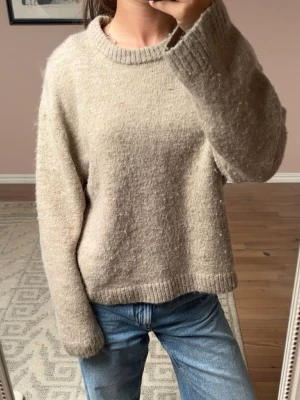Beige stickad tröja i ullmix - Mysig beige stickad tröja med rund hals och avslappnad passform. Tröjan har ribbade muddar vid ärmslut och nederkant, och är tillverkad i en mjuk ullblandning som känns skön mot huden. Perfekt att styla med jeans för en enkel look.