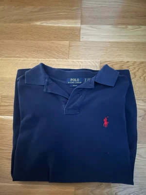 Marinblå pikétröja från Ralph Lauren - Klassisk marinblå pikétröja från Ralph Lauren med korta ärmar och röd broderad logga på bröstet. Tröjan har krage och knäppning framtill. Perfekt för en stilren och avslappnad look. Säljer då den är för liten.