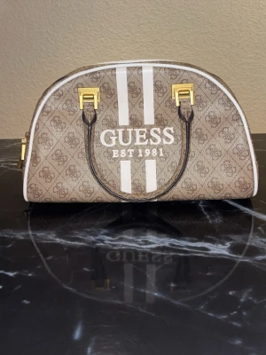Beige handväska från Guess - Snygg beige handväska från Guess med logomönster, vita ränder och guldfärgade detaljer. Väskan har rundad form, dubbla handtag och dragkedja baktill. Materialet är konstläder och väskan har tydlig Guess-logga framtill.