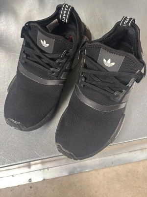 Svarta Adidas NMD_R1 sneakers - Säljer ett par svarta Adidas NMD_R1 sneakers med klassiska tre ränder på sidan och vit Adidas-logga på plösen. Ovandelen är i mesh och syntetmaterial, med svarta snören och detaljer i svart och vitt på hälen. Skorna har en sportig och modern look.
