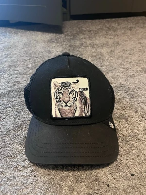 Svart truckerkeps med tigerpatch - Säljer en svart truckerkeps från Goorin Bros med mesh på sidorna och bak. Framtill sitter en broderad patch med en tiger och texten 'TIGER'. Kepsen har böjd skärm och justerbar snapback. Perfekt för dig som vill ha en cool och djurinspirerad stil.