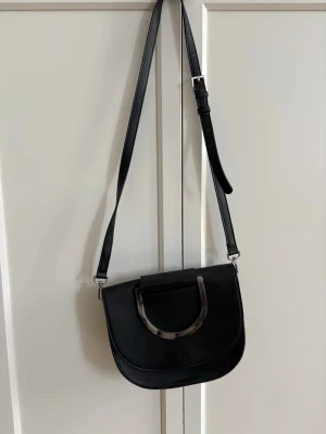 Svart axelväska från Saint Laurent - Snygg svart axelväska från Carin Wester i slätt läder med justerbar axelrem och unik halvcirkelformad detalj i marmorerad plast på locket. Väskan har en stilren och modern design som passar perfekt till din outfit.