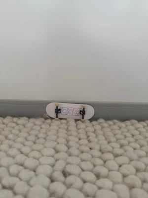 Vit fingerboard med grafik - Säljer en vit fingerboard med färgglad grafik på ovansidan där det står 'OSLID'. Undersidan är helt vit och brädan har små vita hjul och svarta truckar. Perfekt för dig som gillar att göra tricks med fingrarna.