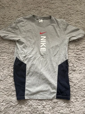 Grå Nike t-shirt med logga - Snygg grå t-shirt från Nike med vit vertikal logga och röd swoosh på bröstet. T-shirten har mörkblå paneler på sidorna och är tillverkad i mjuk bomull. Klassisk passform och korta ärmar, perfekt för en sportig och avslappnad stil.