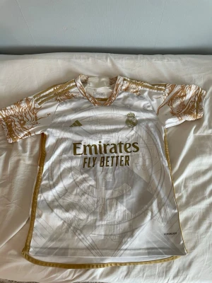 Real Madrid Adidas - Tshirt - (Pris kan diskuteras) Snygg vit Real Madrid t-shirt från Adidas med guldiga detaljer och tryck. På ryggen står det RONALDO och nummer 7 i guld. Framsidan har klubbmärke, Adidas-logga och Emirates Fly Better-tryck. Materialet är polyester och modellen är kortärmad.