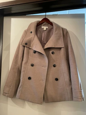 Beige kappa (skepparkavaj) från H&M - Stilren beige kort kappa från H&M med dubbelknäppning och stora svarta knappar. Kappan har bred krage, raka ärmar och är fodrad med matchande tyg. Perfekt för dig som vill ha en klassisk look med modern touch. Väldigt len och clean material. 