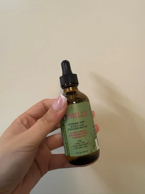 Mielle Rosemary Mint Scalp & Hair Oil - Mielle Rosemary Mint Scalp & Hair Strengthening Oil i en brun glasflaska med pipett. Oljan är berikad med biotin och rosmarin, perfekt för att stärka och vårda hårbotten och håret. Flaskan rymmer 59 ml och har en grön etikett med röda och svarta detaljer. Använd 2 gånger.
