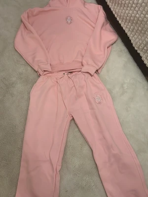Ljusrosa mjukis set från Temps.cb - Säljer ett ljusrosa set från Temps.cb med broderad logga på bröstet. Hoodien har huva, magficka och ribbade muddar vid ärmsluten. Tillverkad i mjuk bomullsblandning som känns skön mot huden. Perfekt för en avslappnad och trendig look. Köpt för 1700kr. Helt oanvänd! Pris kan diskuteras