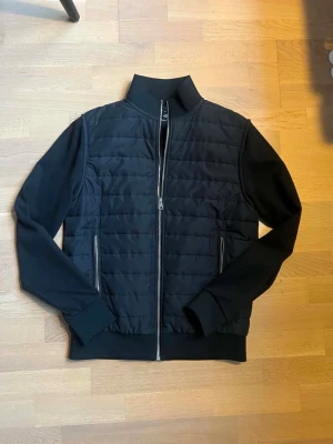 Svart quiltad jacka med dragkedja - Snygg svart jacka med quiltad front och släta ärmar. Jackan har hög krage, två fickor med dragkedja och hel dragkedja framtill. Perfekt för dig som gillar en stilren look med sportig touch.