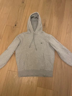 Grå hoodie från Lacoste - Grå Lacoste hoodie i storlek M. Tröjan är i mycket bra skick och har inga defekter. Nypris 1600 kr. Hör av dig vid frågor eller funderingar!