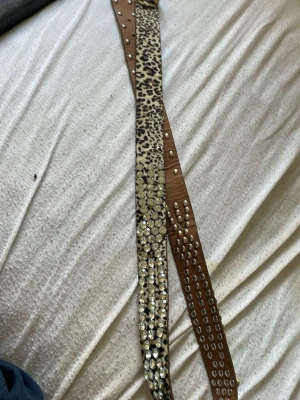Leopardmönstrat nitat bälte - Snyggt bälte med leopardmönster i beige och svart, dekorerat med silvriga nitar längs hela bandet. Spännet är också silvrigt och prytt med små nitar för extra bling. Perfekt accessoar för att lyfta vilken outfit som helst.