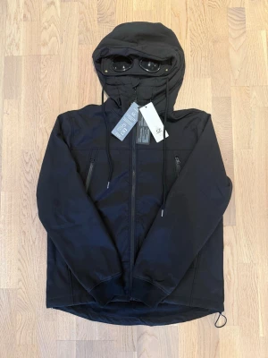 Svart hoodie från C.P. Company - Snygg svart hoodie från C.P. Company med dragkedja framtill och två sidofickor med dragkedja. Huvan har inbyggda glasögon och justerbara snören. Tillverkad i ett mjukt och slitstarkt material som känns riktigt skönt mot huden. Perfekt för dig som gillar streetwear och coola detaljer.
