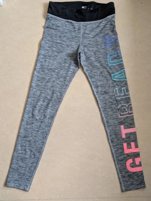 Grå leggings från H&M med tryck - Säljer ett par gråmelerade leggings från H&M med svart midja och färgglatt tryck längs ena benet där det står 'GET READY' i rosa, blått och turkost. Perfekta för träning eller chill. Materialet är stretchigt och mjukt. HELT OANVÄNDA leggings 