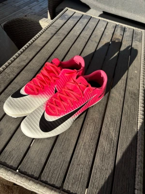 Nike rosa och vita fotbollsskor - Säljer ett par snygga fotbollsskor från Nike i en grymt fräsch rosa och vit färgkombination med svarta detaljer. Skorna har klassisk snörning och är tillverkade i syntetmaterial för bästa känsla på planen. Perfekta för dig som vill sticka ut på matchen!
