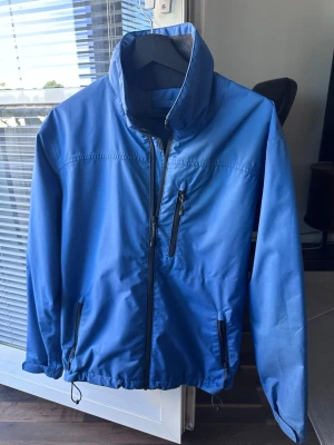 Blå vindjacka från Helly Hansen - Snygg blå vindjacka från Helly Hansen med hög krage och dragkedja framtill. Jackan har tre fickor med dragkedja och justerbar nederkant. Insidan är fodrad med mjuk fleece för extra komfort. Perfekt för blåsiga dagar och outdoor-aktiviteter.