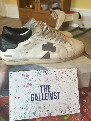 The Gallerist Skor - Säljer dessa snygga low-top sneakers från The Gallerist – ett distinkt märke med konstnärlig touch. Skorna har ett klassiskt silhuett i vitt läder med detaljer i grått och svart, och pryds av ett coolt klöveremblem på sidan. ✔️ Äkta läder ✔️ Märket synligt på tungans överkant ✔️ Säljs med originalkartong ✔️ Använt skick – har patina och karaktär