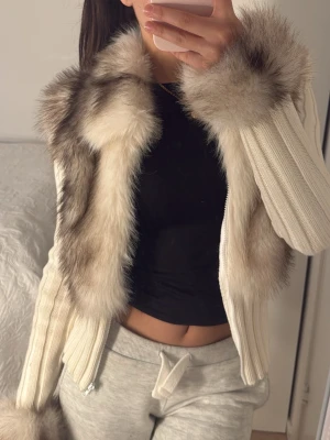 Alessa päls kofta - Säljer en superfin vit pälsjacka med fluffig fuskpäls i beige och grå toner. Jackan har ribbstickade ärmar och dragkedja framtill. Pälsen går längs kragen och ner över framsidan samt vid ärmsluten för extra lyxig känsla.